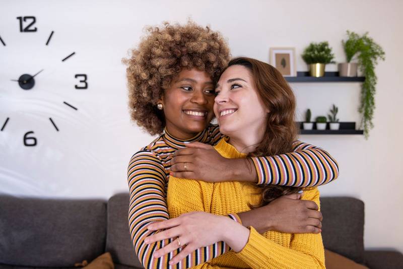 Beautiful and cheerful multiracial lesbian couple hugging and looking each other smiling. Lovely and happy homosexual women embracing at home staring each other.