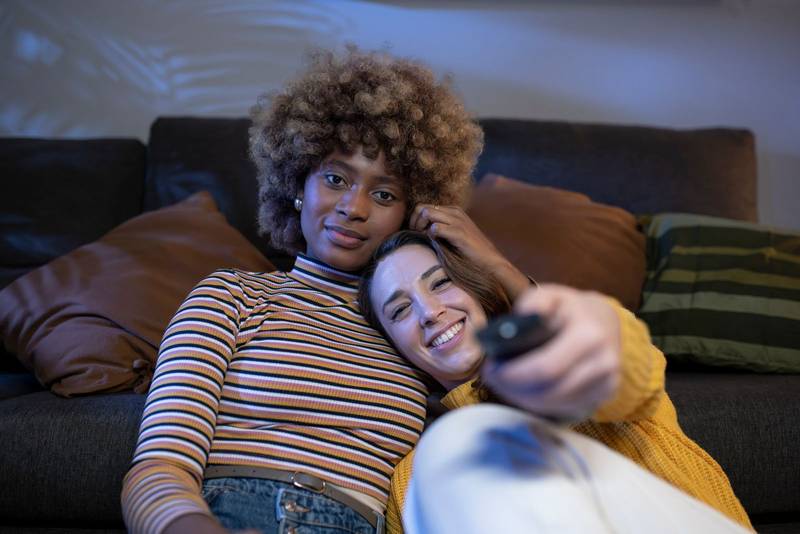 Lesbian multiracial lesbian women looking at camera and using the remote control. Lovely couple relaxing on the floor of a living room smiling and hugging. Focus on girls.
