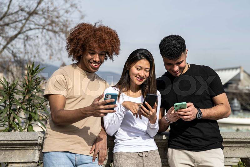 Three young multiracial people texting with smartphones standing in a park. Diverse group smiling and using phones outdoors on a stone balcony