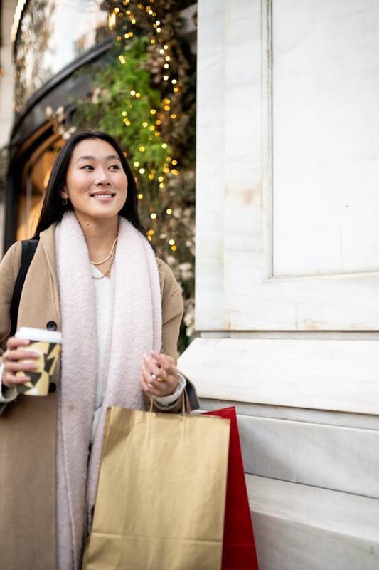 Young carefree woman shopping Christmas presents in the street. Happy joyful girl carrying bags and a coffee in winter.