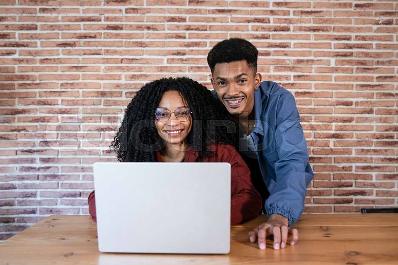Satisfied friends looking at camera and using a computer at cozy home office. Diverse young couple using laptop together at home.