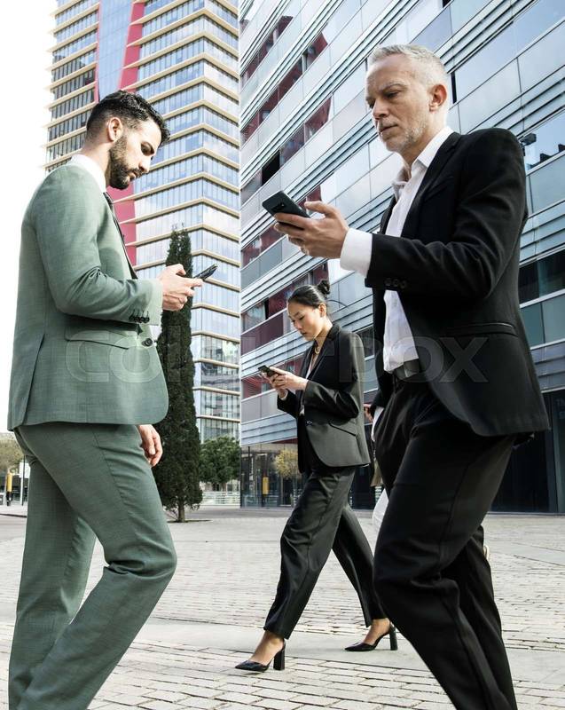 Side view of a group of businessmen and a businesswoman walking in the street while looking at their phones. Multiracial businesspeople checking their phones while waking in the city.