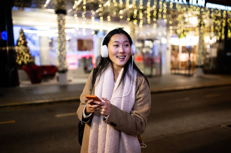 Young relaxed girl texting and wearing headphones with christmas lights behind. Happy woman listening to music at the street during winter using her phone.